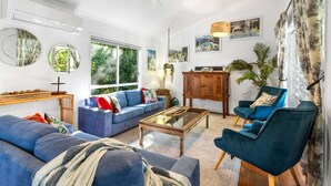 A spacious living room with a blue sofa, a wooden coffee table, and a blue armchair. There is a large window providing natural light and a view of greenery outside. - Tropical Family Hideaway, Noosa Heads (Noosa Heads)