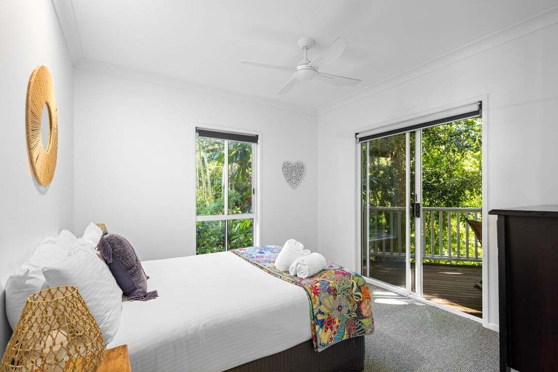 Un dormitorio con cama, ventilador de techo, decoración con forma de corazón y ventanas amplias con vista a la vegetación.