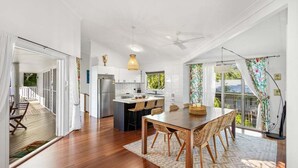 A modern kitchen with white cabinets, a central island, and a dining area with a wooden table and woven chairs. - Tropical Family Hideaway, Noosa Heads (Noosa Heads)