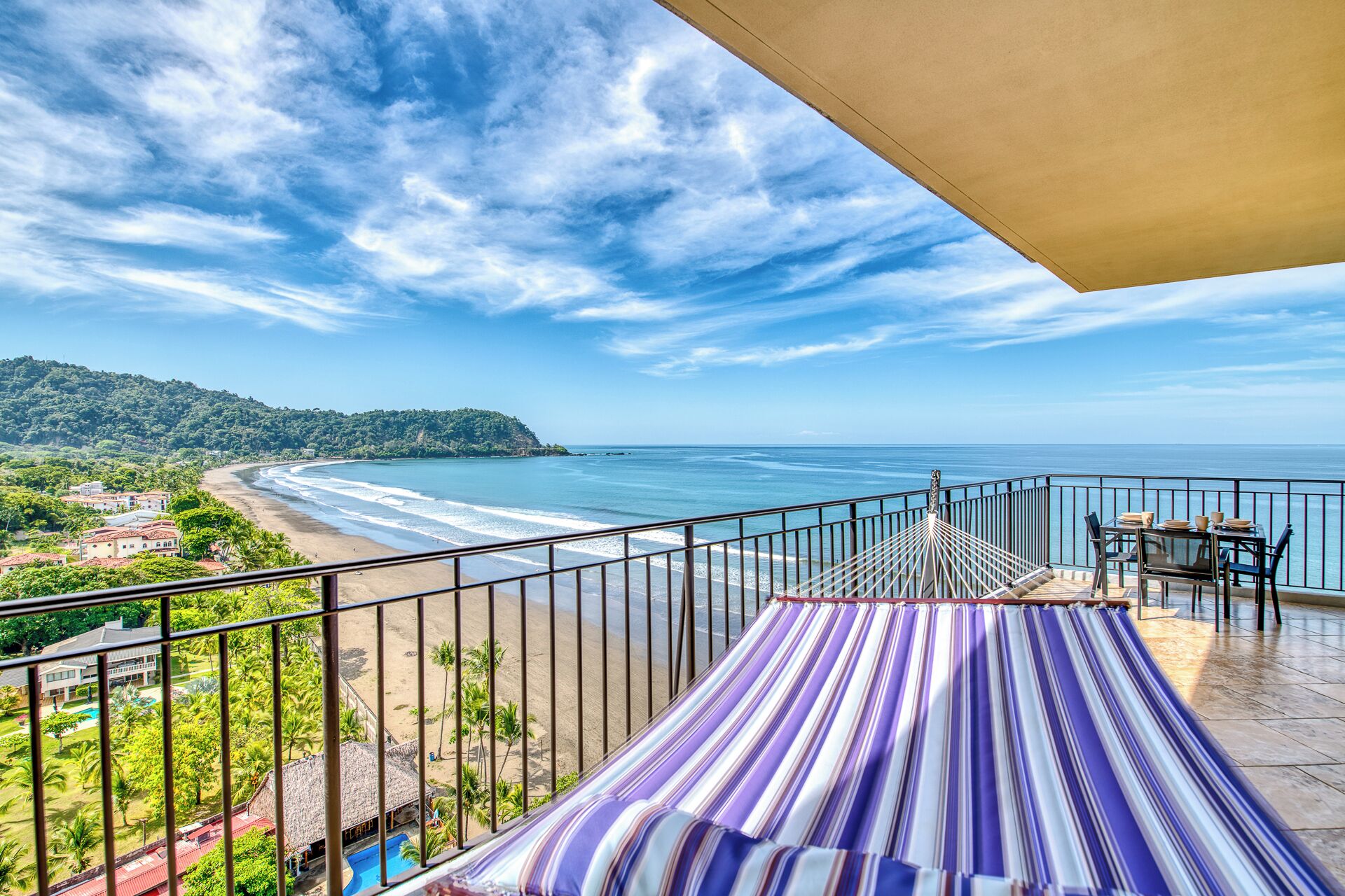 A balcony with a view of a beach and ocean, a hammock, and a table with chairs.