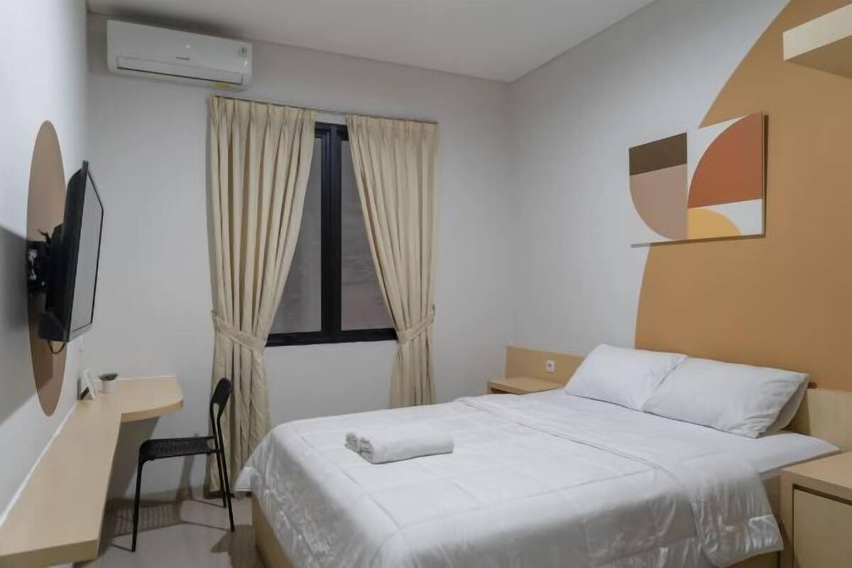 A hotel room with a bed, a desk, a chair, a TV, and a window with curtains.