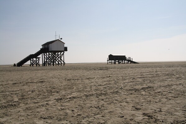 A beach with two wooden structures, one with a staircase and another with a flat roof. - Ferienhaus in St. Peter-ording (St. Peter-Ording)