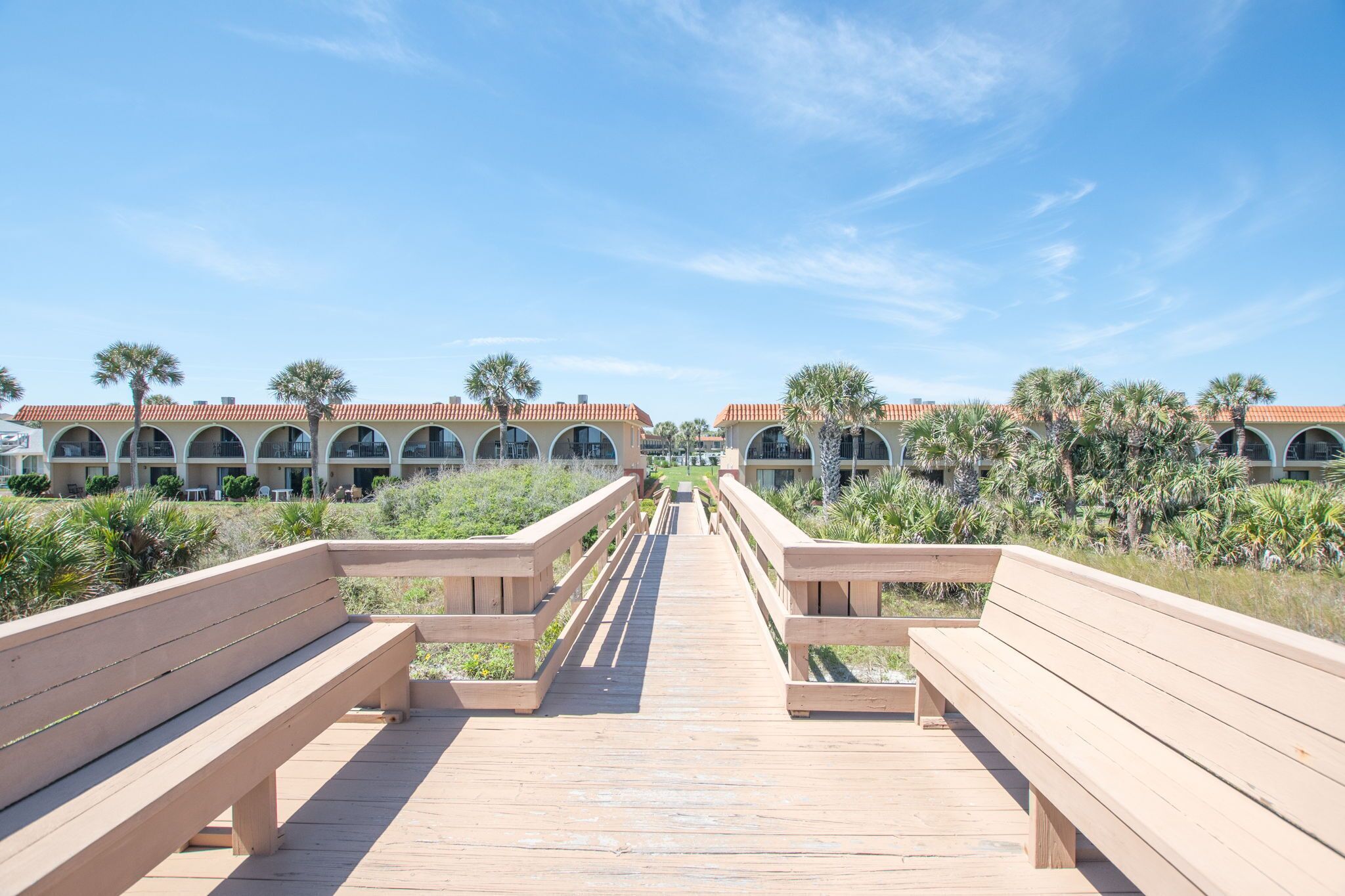Un paseo marítimo de madera que conduce a una hilera de edificios con ventanas arqueadas bajo un cielo azul despejado.