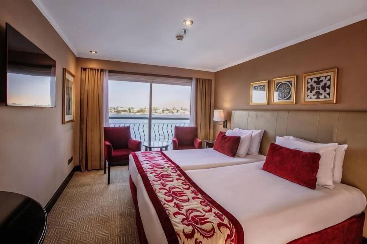 Ein Hotelzimmer mit zwei Betten, einem Fernseher, einem Schreibtisch, einem Sessel und einem Balkon mit Blick.