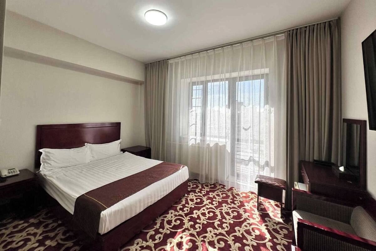 Een hotelkamer met een groot bed, een nachtkastje met een telefoon, een televisie aan de muur en uitzicht op een stadsgezicht door dunne gordijnen.