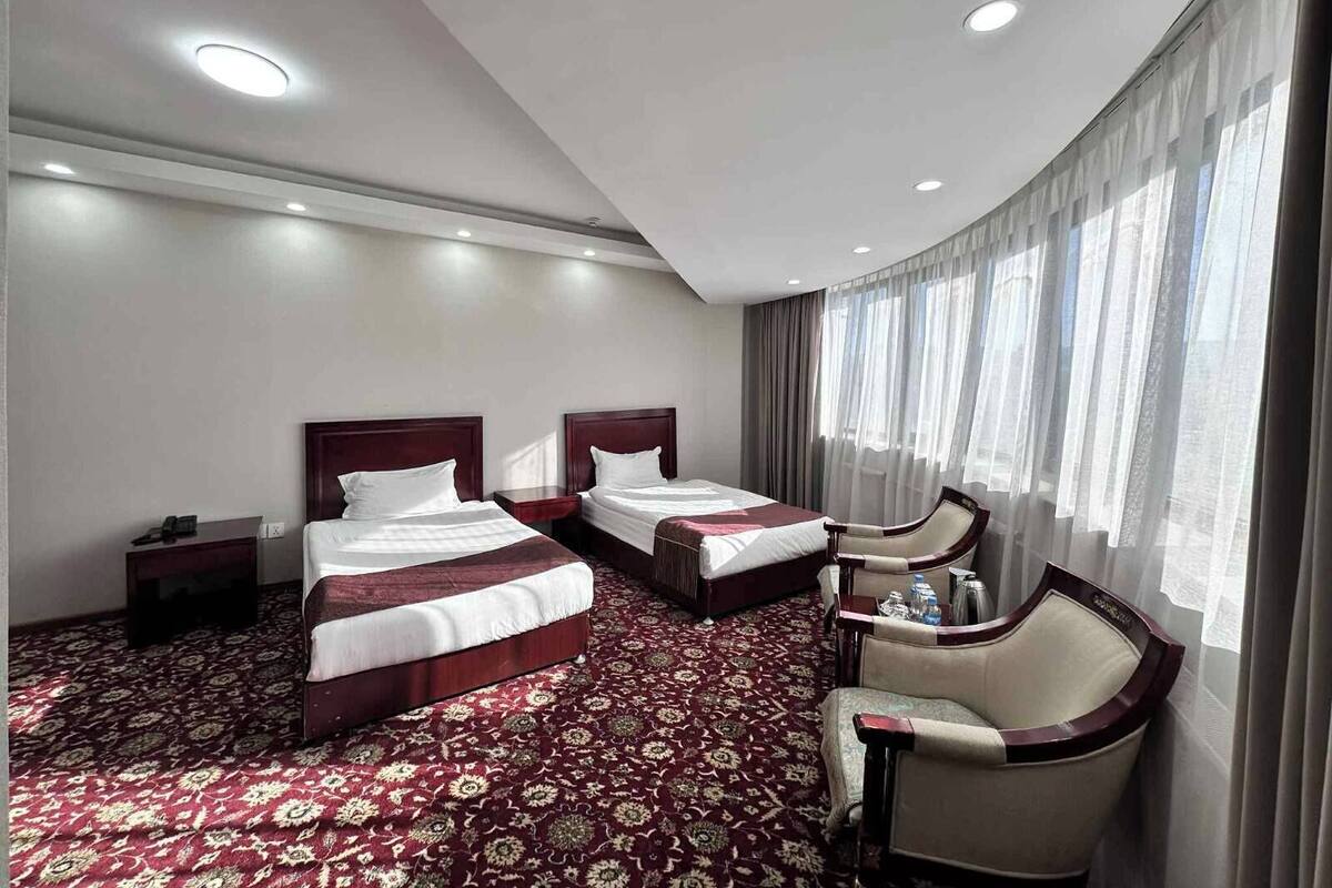 Een hotelkamer met twee bedden, een zithoek met een stoel, en een groot raam met gordijnen.