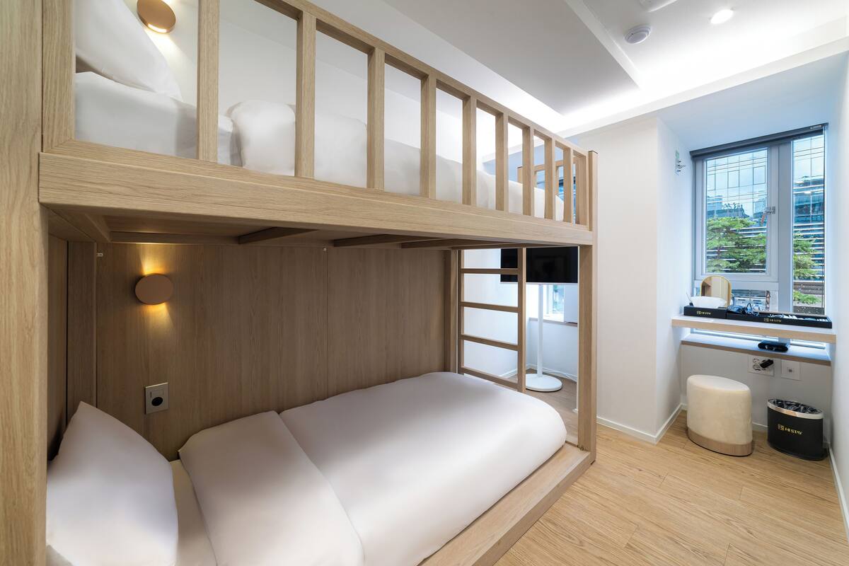 Una cama literera con estructura de madera, una mesita de noche y una ventana con vistas a zonas verdes.
