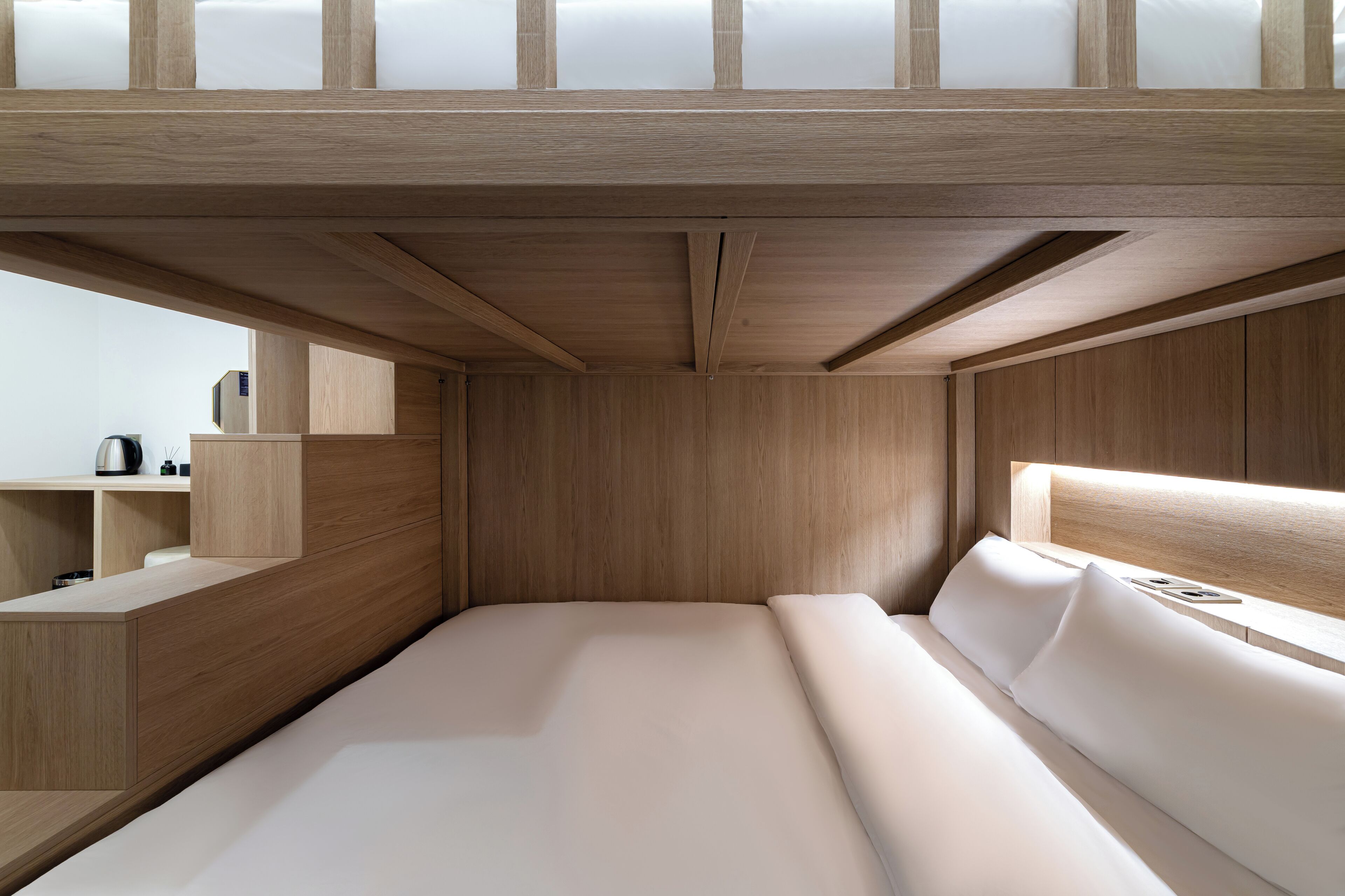 A modern, minimalist bedroom with a wooden headboard, a neatly made bed, and a built-in desk with a kettle.
