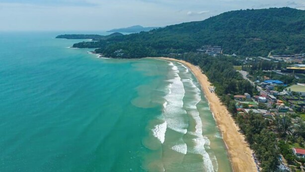Oversigtsbillede af et kystområde med strand, bygninger og en skovklædt bakke.