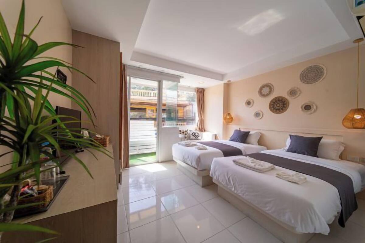 A hotel room with two beds, a large potted plant, a balcony with a view, and decorative wall plates.