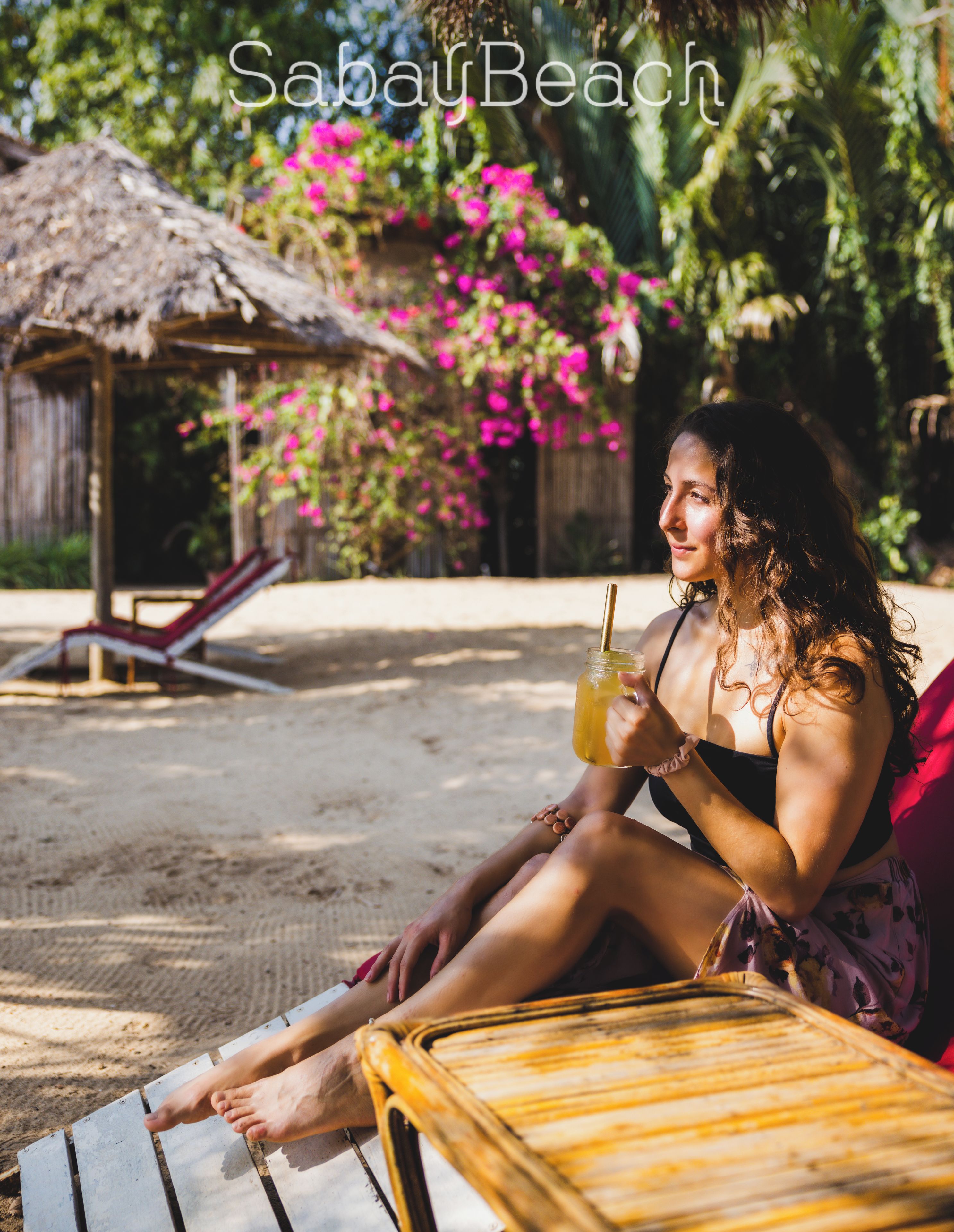 A person relaxing on a wooden lounge chair with a drink, a thatched-roof hut, and pink flowers in the background.