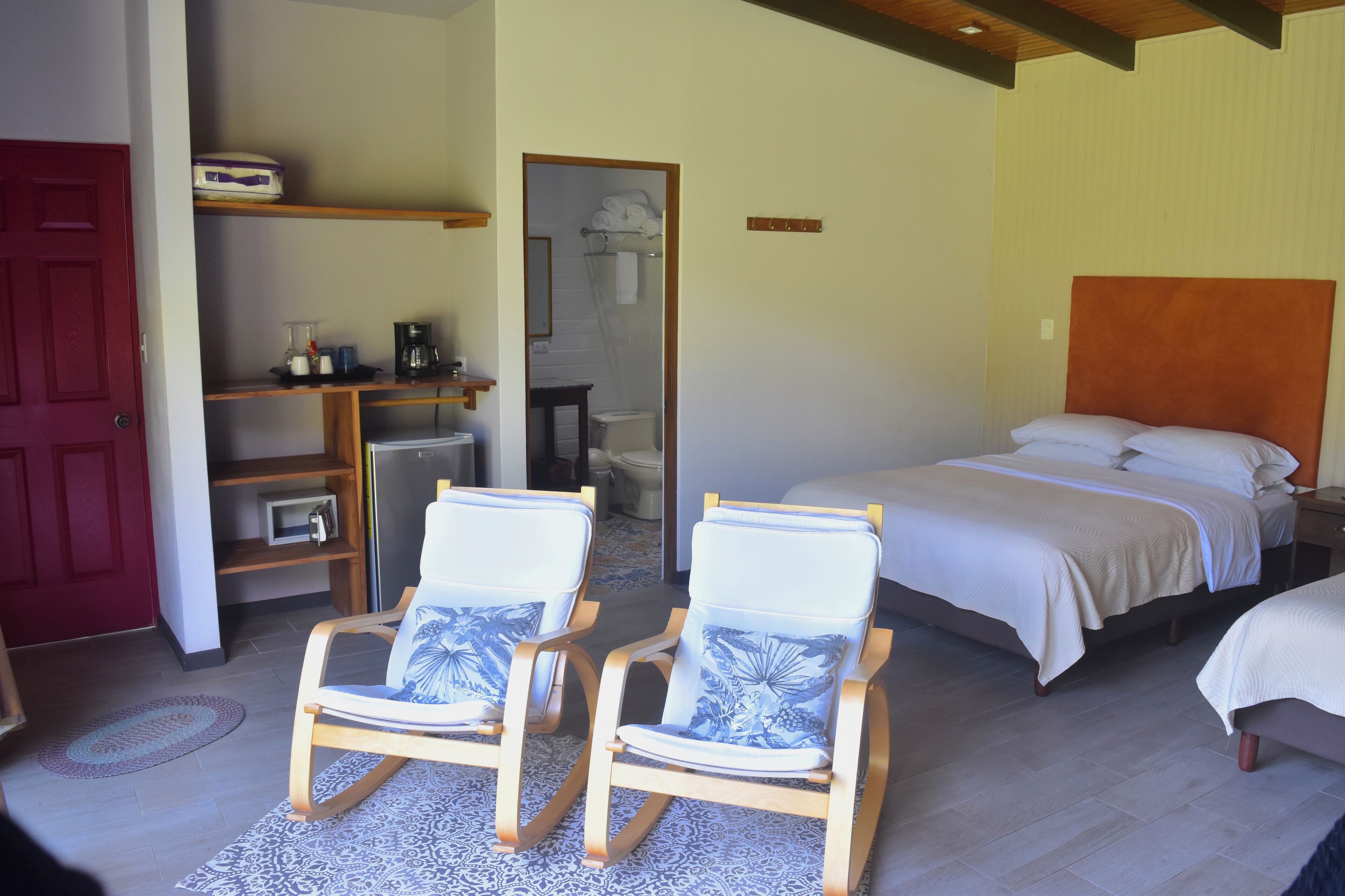 A bedroom with a bed, two rocking chairs, a desk with a coffee maker, and a red door.