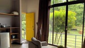 Una habitación con sofá, una mesita con planta, zona de cocina con refrigerador y microondas, y un ventanal con vistas a zonas verdes.