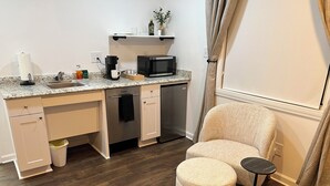 Cocina compacta con microondas, cafetera y lavabo. Hay un taburete pequeño y un otoman redondo en la habitación.