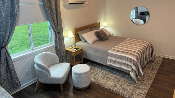 Un dormitorio con cama, lámparas de noche, una silla, un reposapiés, una ventana con cortinas, un equipo de aire acondicionado y un espejo en la pared.