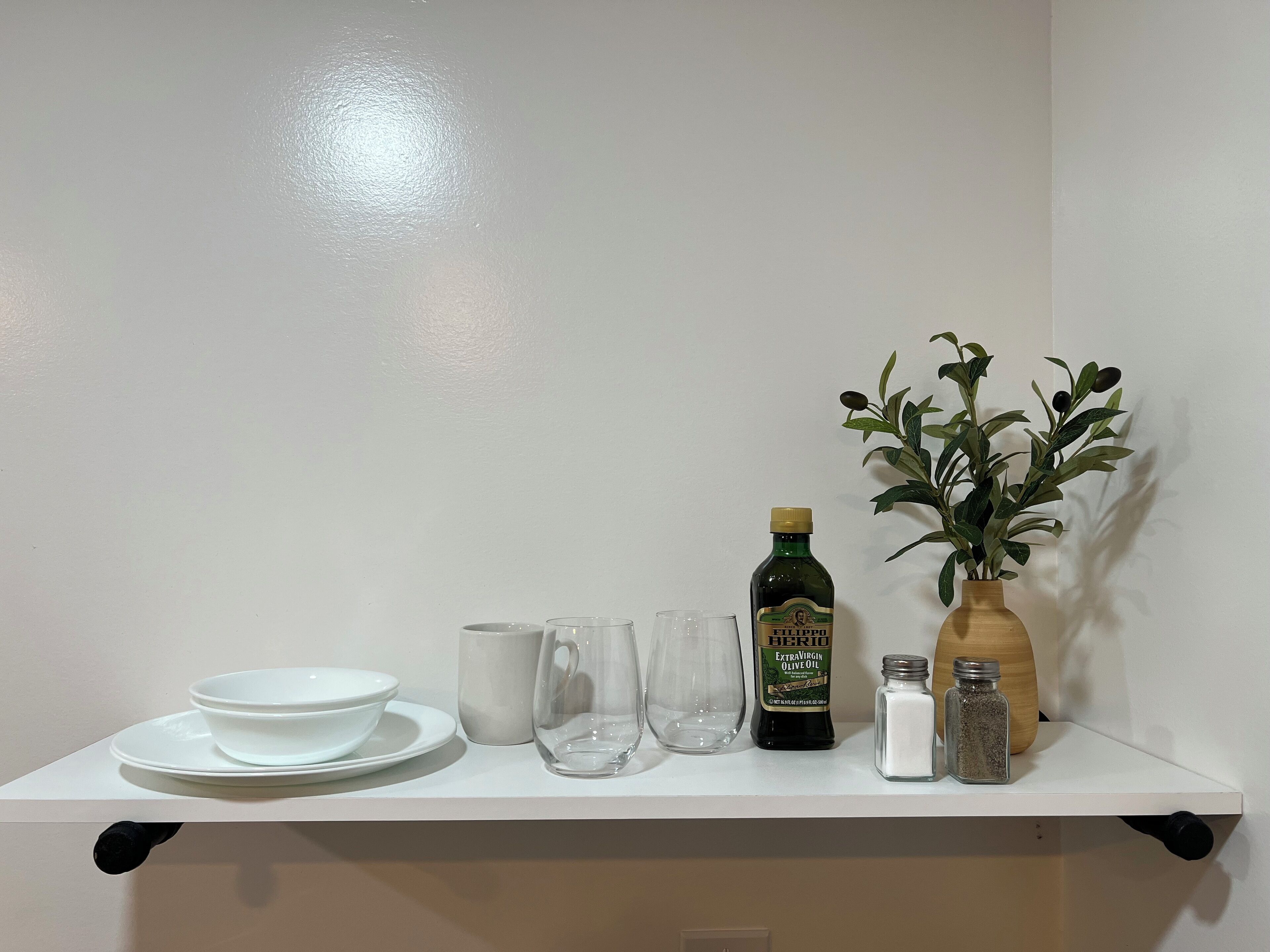 A white shelf with a bottle of olive oil, a vase with green leaves, salt and pepper shakers, and a stack of white plates and bowls.