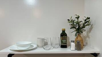 A white shelf with a bottle of olive oil, a vase with green leaves, salt and pepper shakers, and a stack of white plates and bowls.
