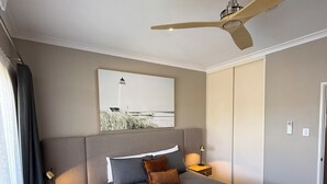 Una habitación de hotel moderna con una cama grande, un ventilador de techo, una mesita de noche con lámpara y un cuadro de un faro en la pared.