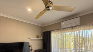 Habitación de hotel con cama, televisor sobre un mueble de madera, ventilador de techo y ventana con cortinas translúcidas.