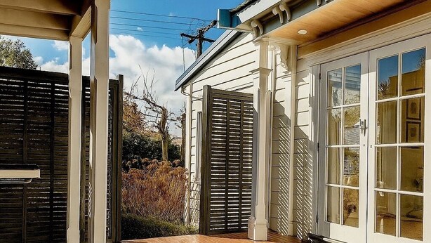 A covered patio with wooden flooring, a white exterior wall, and a sliding glass door leading to a garden.