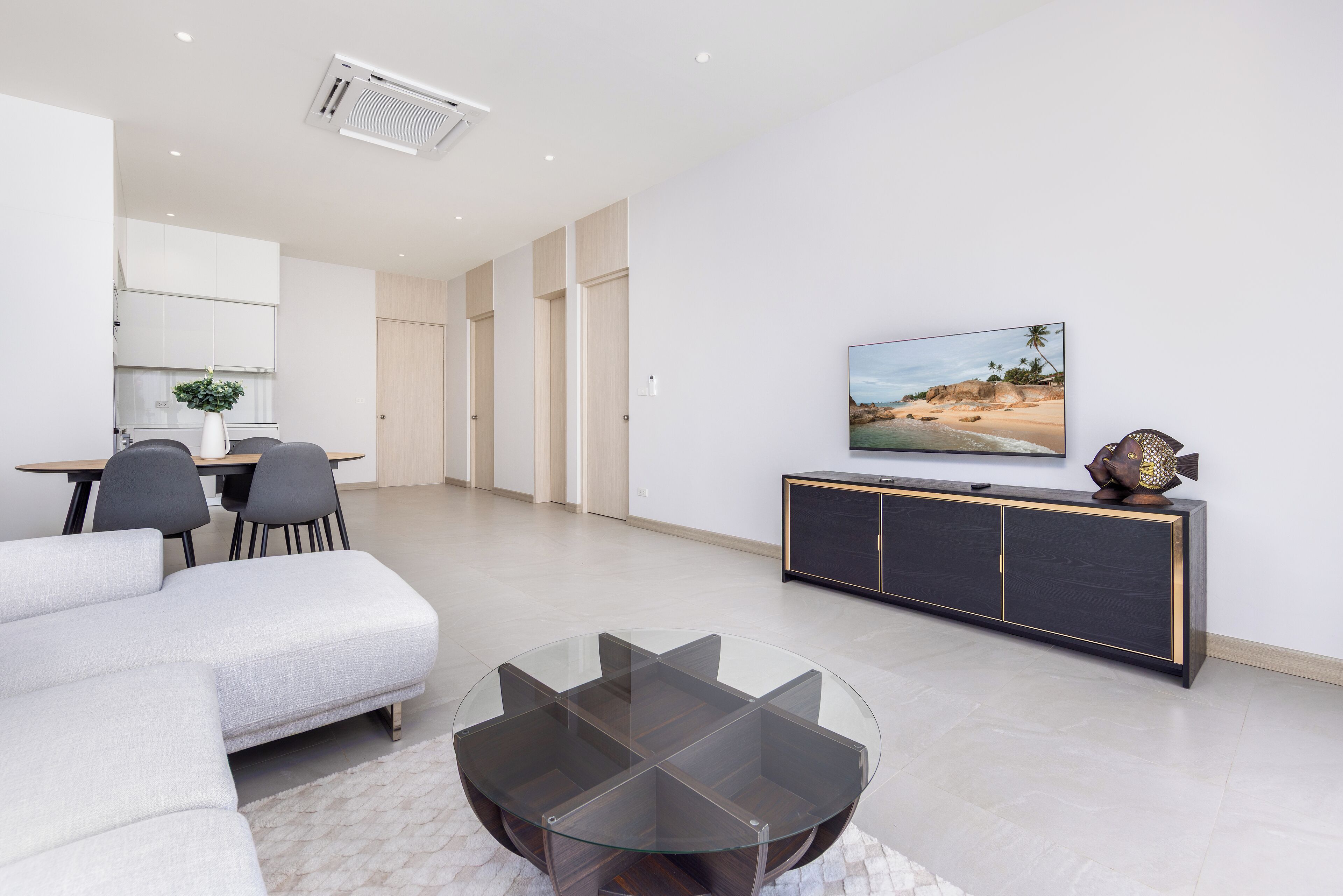 Een moderne woonkamer met een hoekbank, een glazen salontafel, een eethoek met tafel en stoelen, een televisie op een zwart kastje, en een strandtafereel aan de muur.