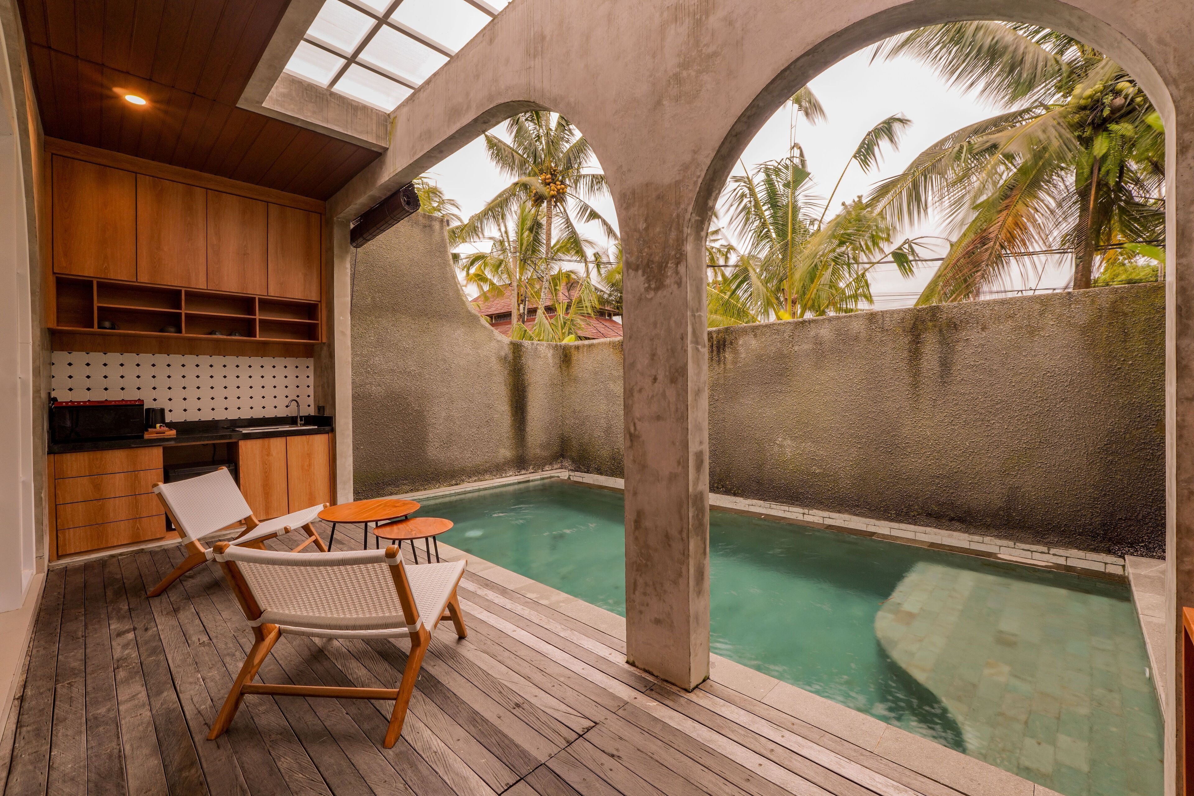 A pool area with wooden deck, lounge chairs, and a kitchen area with wooden cabinets.