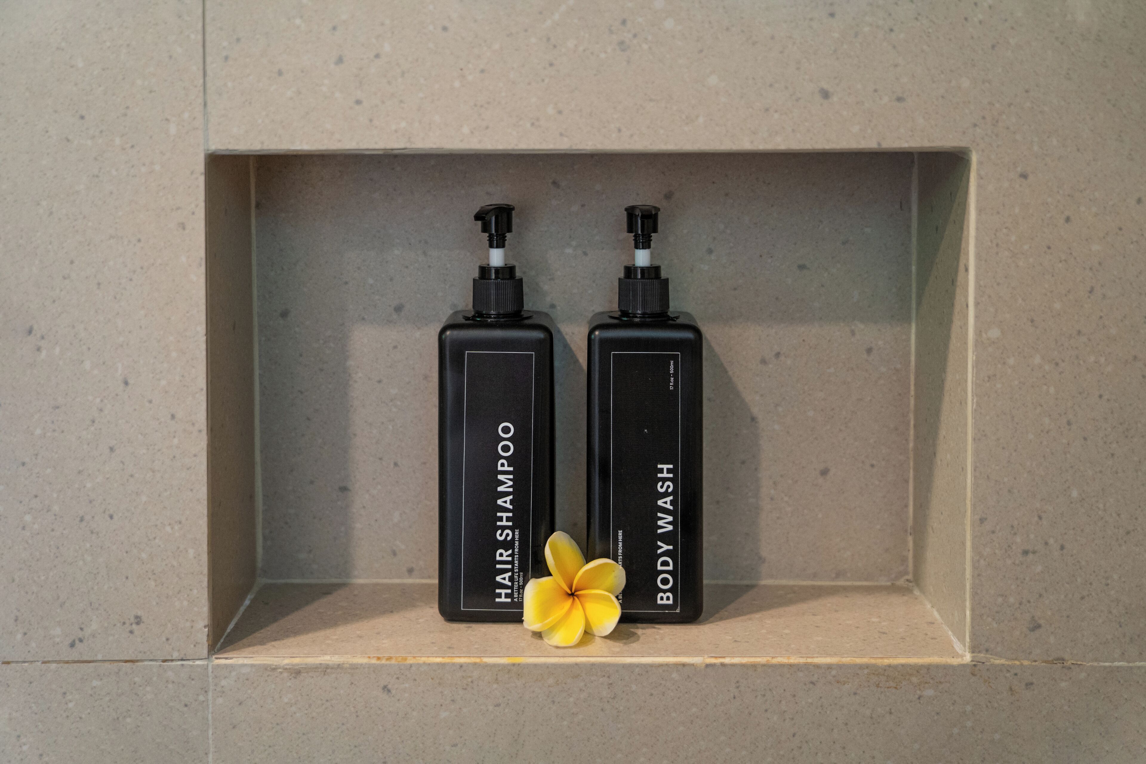 Zwei schwarze Flaschen mit den Aufschriften 'Hair Shampoo' und 'Body Wash' auf einem Fliesenregal.