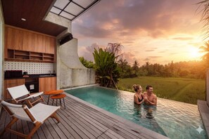 A pool area with lounge chairs, a wooden deck, and a view of a sunset over a rice field. - Subu Utamas Villas (Payangan)