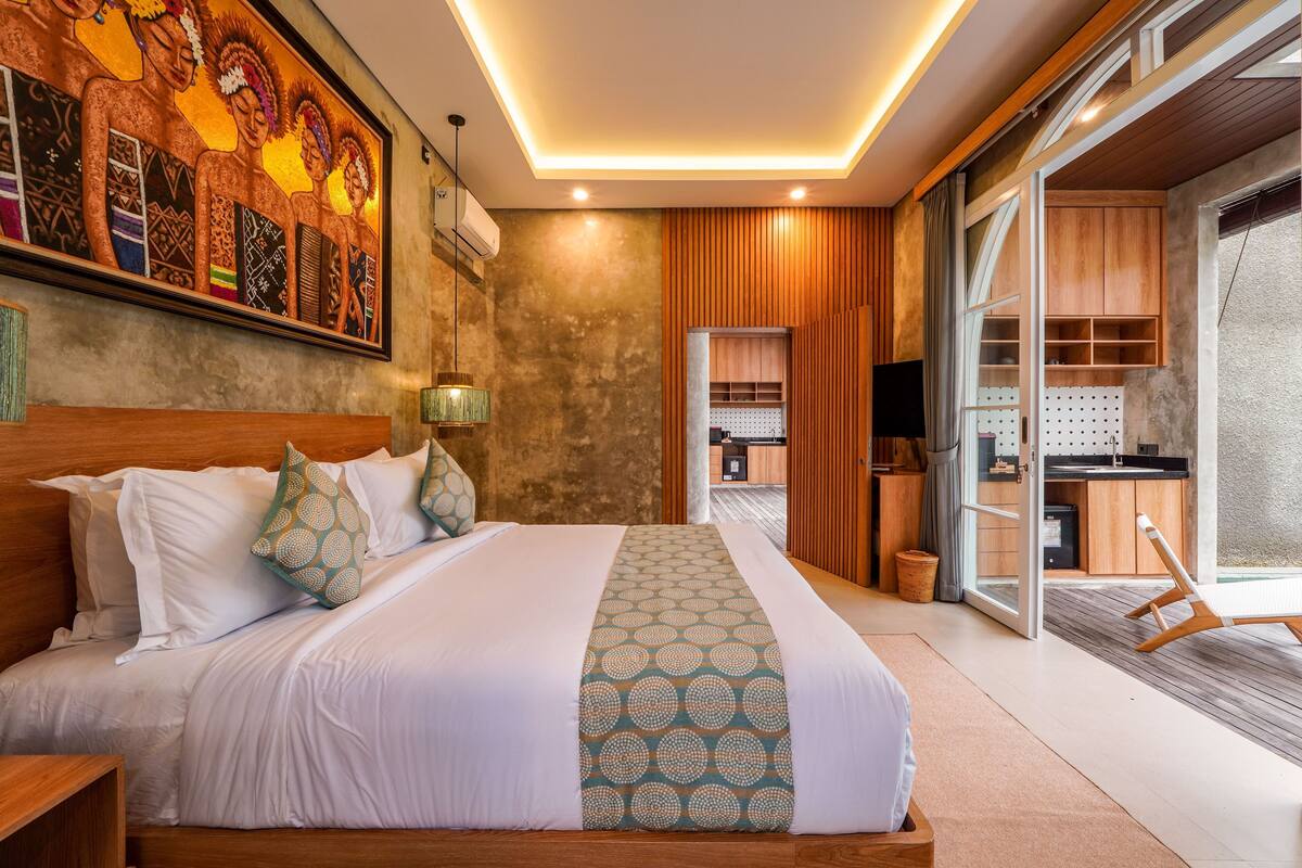 A modern hotel room with a large bed, a painting on the wall, a kitchen area, and a sliding glass door leading to a balcony.