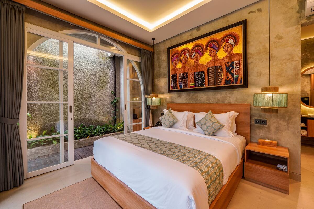 A bedroom with a large bed, a sliding glass door, a painting on the wall, and a nightstand with a lamp.
