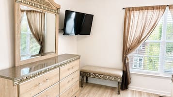Une chambre à coucher avec une commode en bois, un banc, un téléviseur à écran plat fixé au mur et une fenêtre avec des rideaux.
