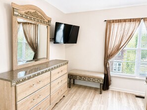 Un dormitorio con un aparador de madera, un banco, un televisor de pantalla plana montado en la pared y una ventana con cortinas.