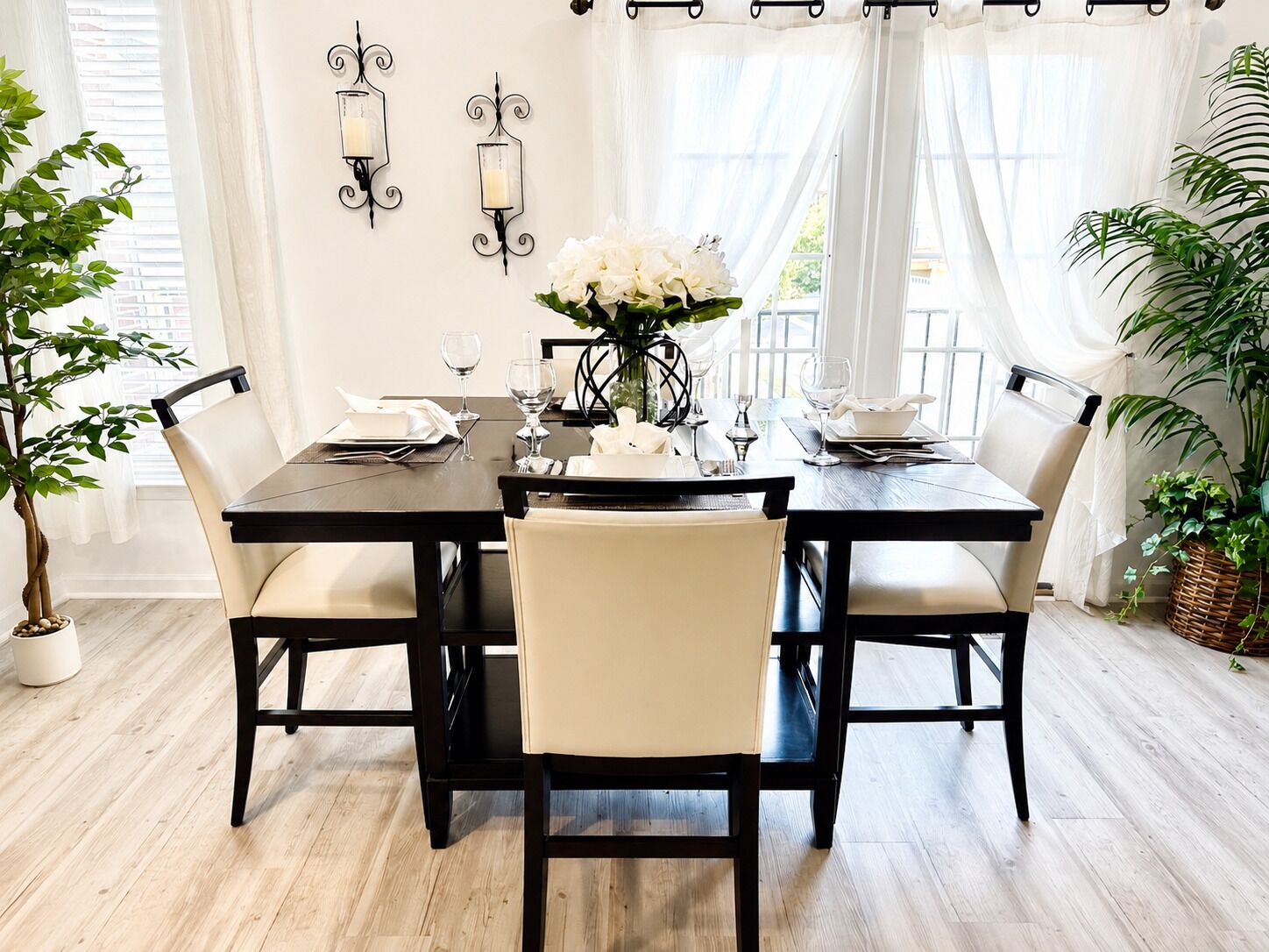 Un comedor con una mesa de madera oscura, sillas blancas acolchadas y un jarrón con flores blancas.