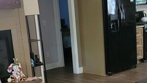 Un salón amplio con suelo de madera, un refrigerador negro y una cocina al fondo.