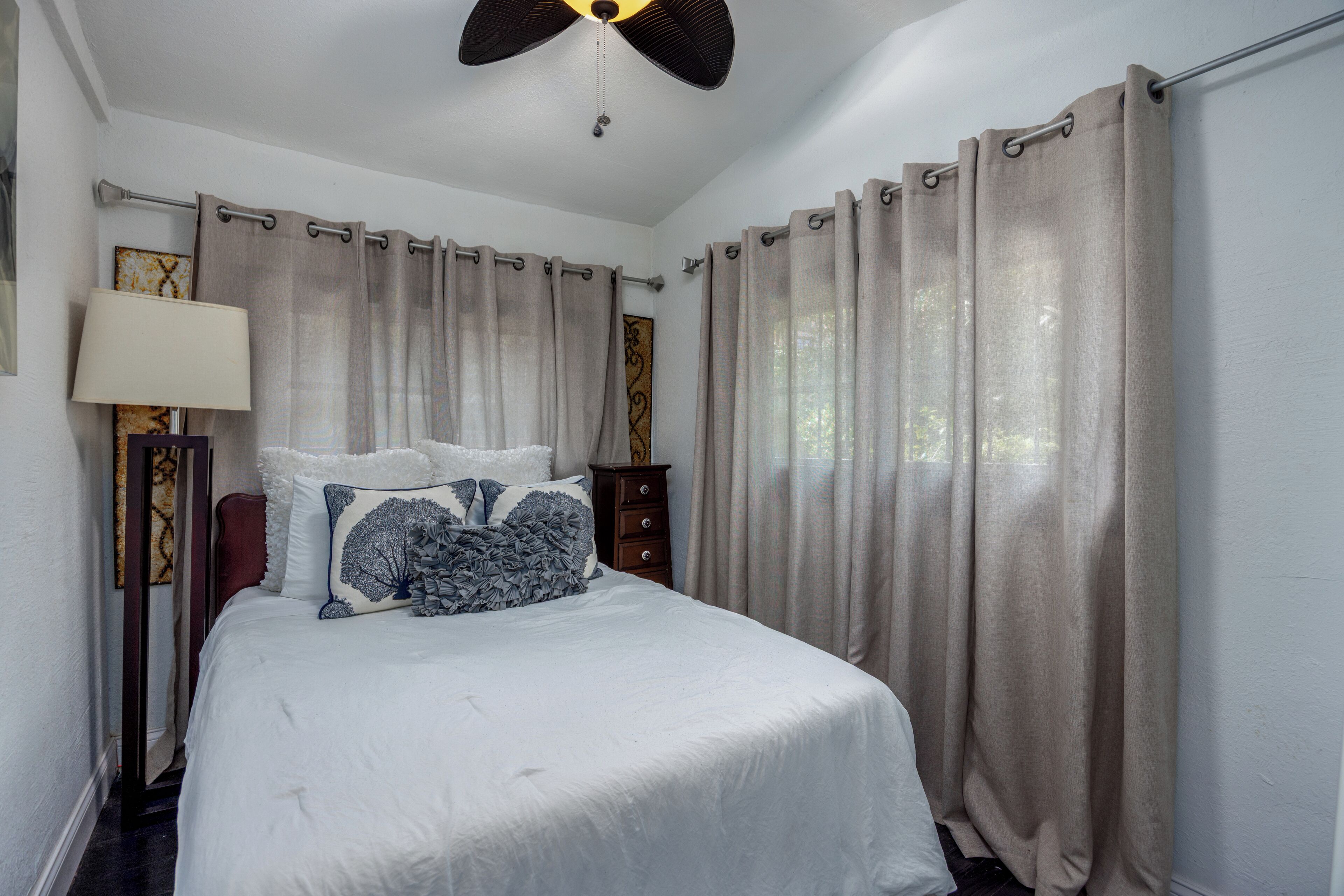 A bedroom with a bed, curtains, a ceiling fan, a nightstand, and a lamp.