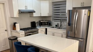 A modern kitchen with white cabinets, a stainless steel refrigerator, a microwave, and a gas stove. There are two blue bar stools at the island.