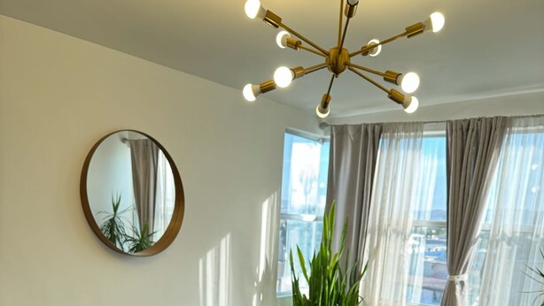 A modern living room with a sectional sofa, a round mirror, a ceiling light fixture, and large windows with curtains.