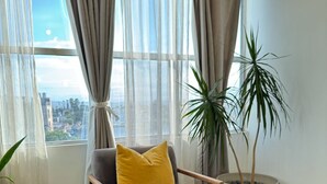 A room with a view of the city, a grey chair with a yellow pillow, a large potted plant, and a teal sofa.