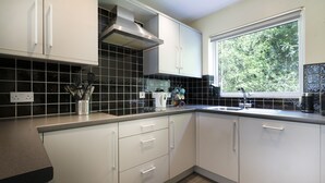A modern kitchen with white cabinets, black tiled backsplash, and a window providing a view of greenery outside. - 22 Thrang Brow -  3 Bedroom Holiday Home - Langdale Valley (Chapel Stile)