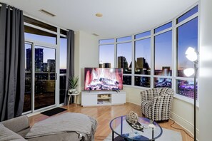 Uma sala de estar moderna com uma televisão grande, um sofá, uma poltrona estampada e uma mesa de centro de vidro.