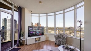 Ett modernt vardagsrum med en stor TV, en soffa, ett soffbord och utsikt över stadsbilden genom stora fönster.