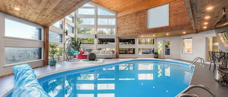An indoor swimming pool with wooden ceiling beams, large windows, and modern furniture.