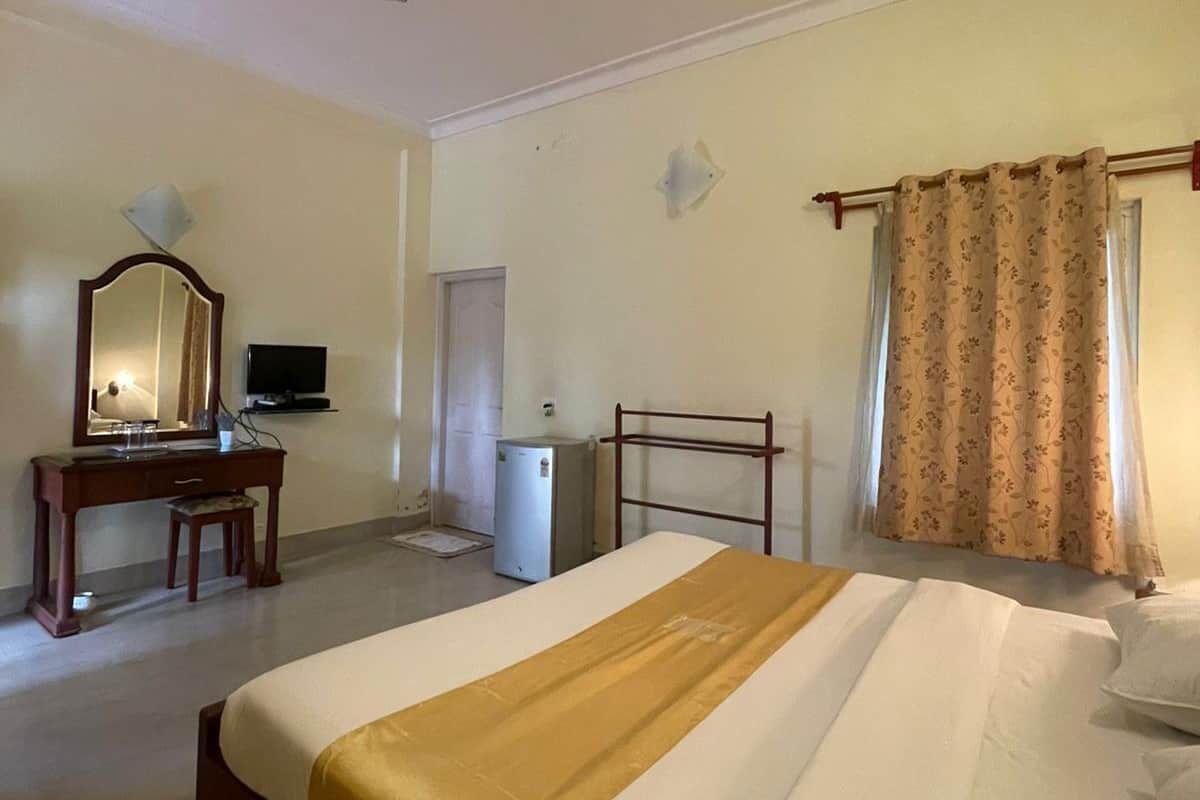 A hotel room with a single bed, a desk with a chair, a television, a ceiling fan, and a window with curtains.