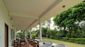 A covered outdoor area with a table and chairs, a glass-top table, and a view of a garden with trees and bushes. - The Grand Oak By TERASTAY (Virajpet)