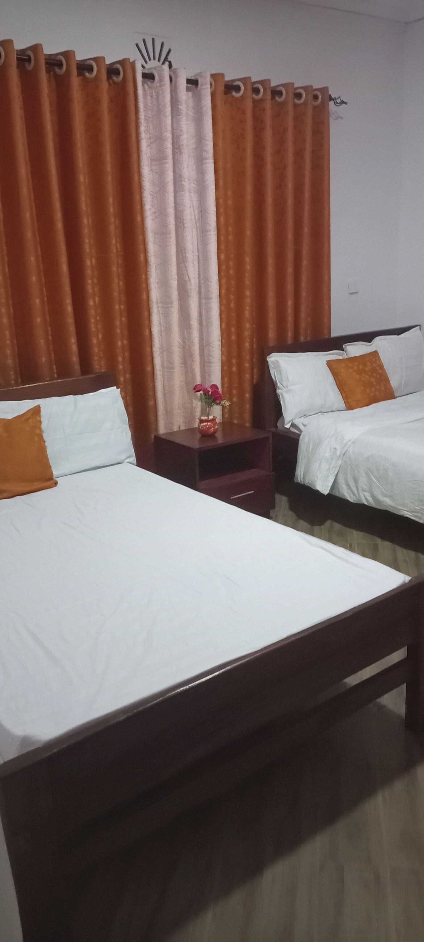 A hotel room with two beds, a nightstand with a vase of flowers, and curtains.