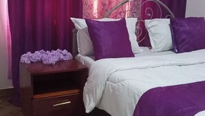 A bedroom with a bed, purple and white bedding, a nightstand with a drawer, a white rug, and purple curtains. - Lux Suites Siyoi Gardens Kitale (Kitale)