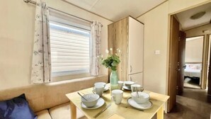 Una habitación con una mesa de comedor lista para cuatro personas, un sofá con un cojín azul, una ventana con cortinas y un armario de madera.