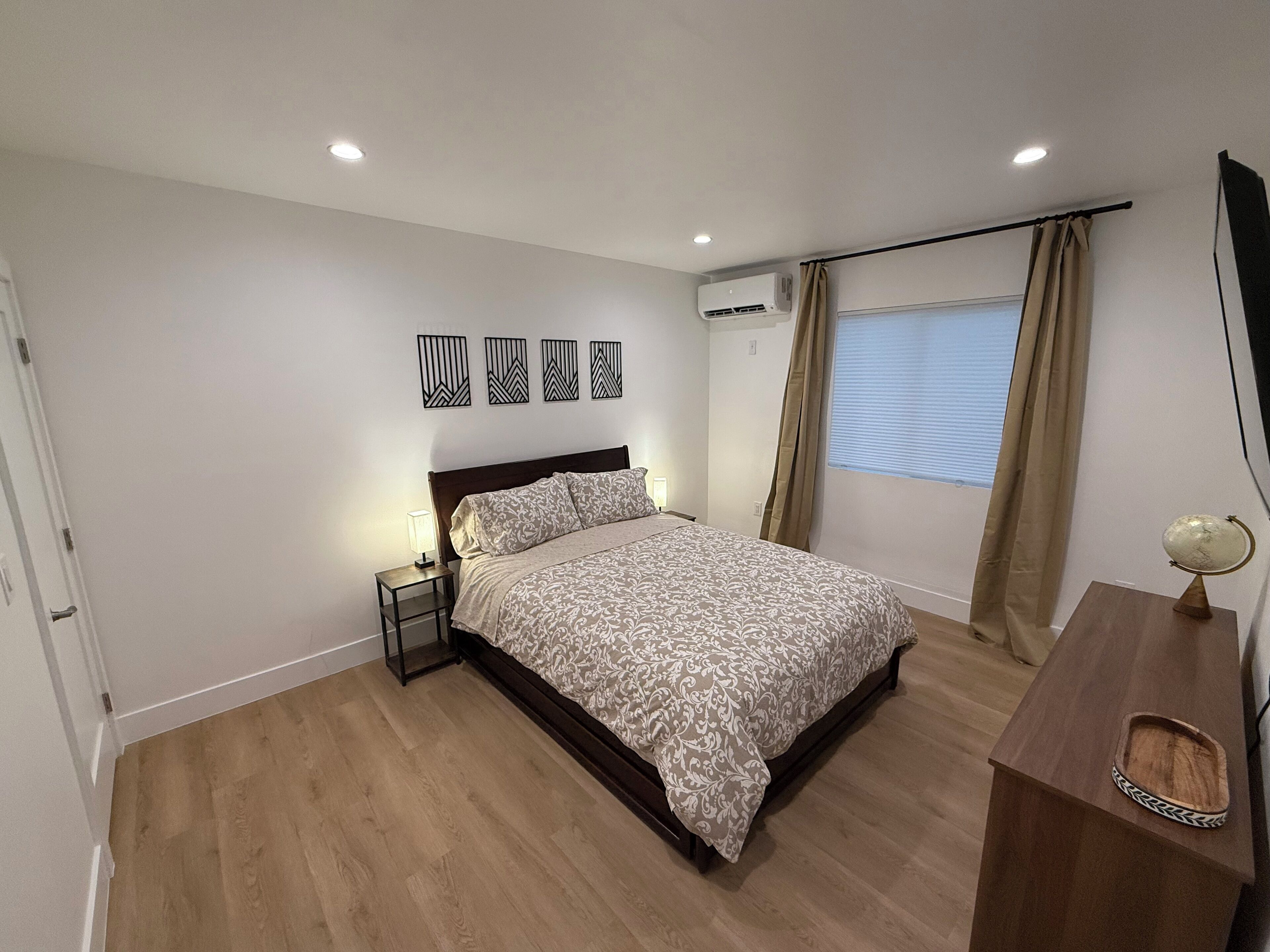 A bedroom with a bed, bedside table, lamp, air conditioning unit, window with curtains, and a wooden dresser.