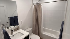 Moderni kylpyhuone, jossa on valkoinen pesuallas, tummat kaapit, wc-istuin, beigenvärinen suihkuverho ja suihku sekä kylpyamme.