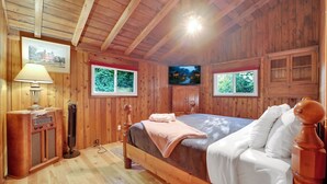 Ein Zimmer mit Holzboden, einem Bett, einer Lampe, einem Fernseher und einem Fenster.